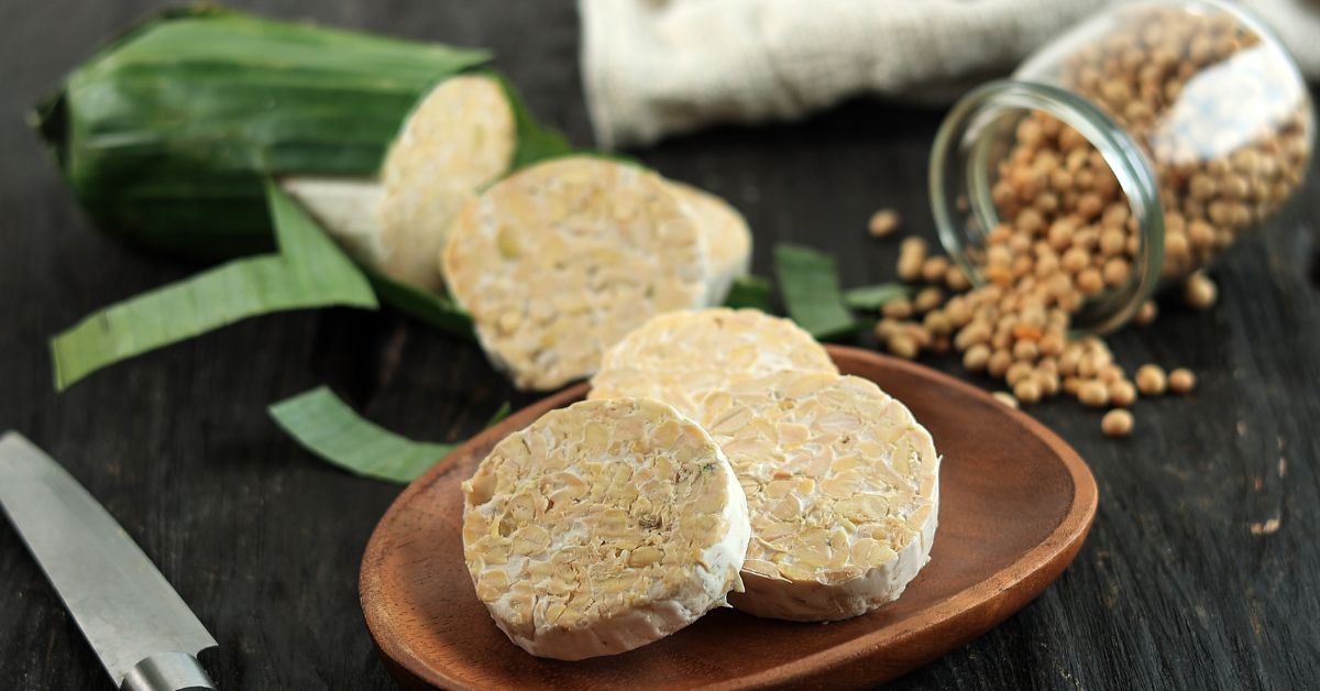 Sekelompok kedelai yang sudah difermentasi menjadi tempe, dibungkus rapi dalam daun pisang, diletakkan di atas meja kayu dengan latar belakang dapur tradisional Indonesia.